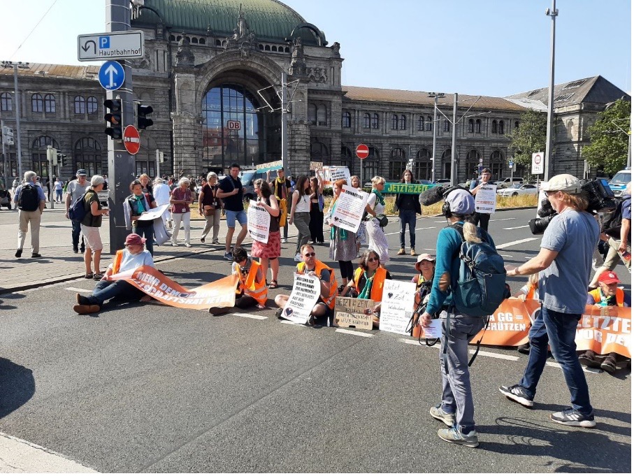 Auf dem Bild ist eine Straße zu sehen, auf der Menschen mit Schildern sitzen. Im Hintergrund ist der Hauptbahnhof zu sehen.
