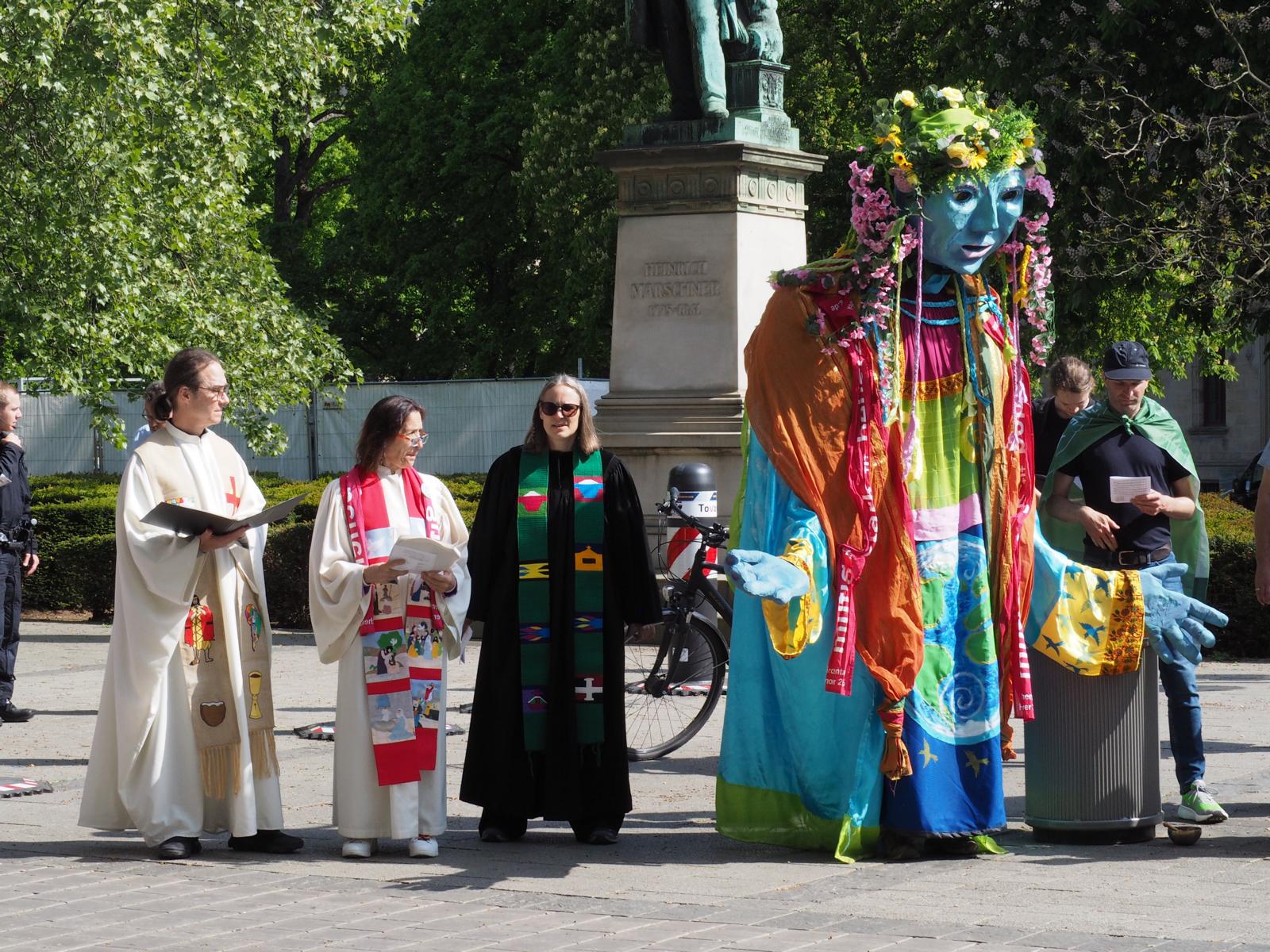 Die drei Pfarrer*innen im Talar und die Gaia-Figur beim Protestmarsch