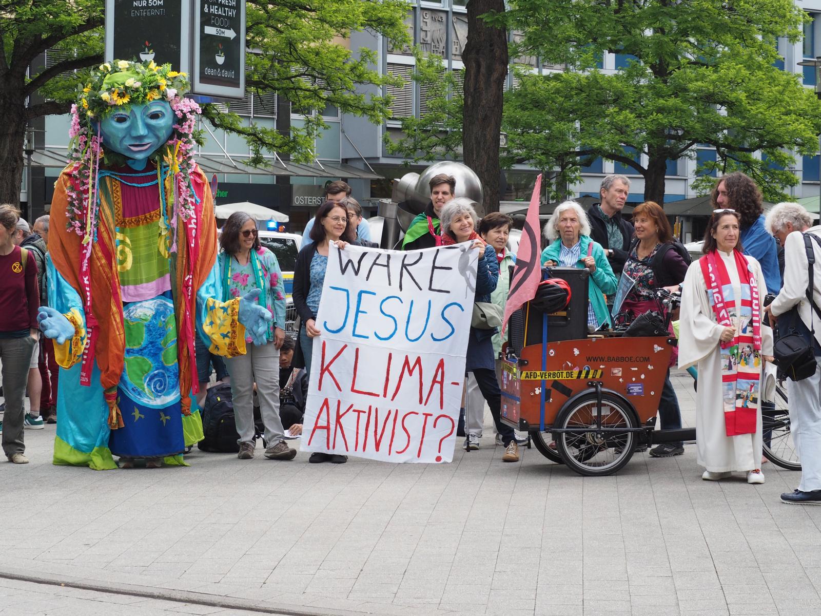 Die Gaia-Figur und fröhliche Menschen neben dem Banner "Wäre Jesus Klimaaktivist?"