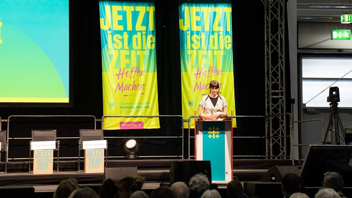 Das Bild zeigt Carla Hinrichs auf einer Bühne, an einem Redepult. Im Vordergrund ist Publikum zu sehen.